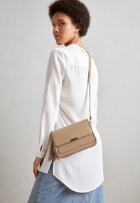 Borsa a tracolla beige realizzata in materiale liscio, con chiusura a pattina, dettaglio con frange e una lunga tracolla regolabile.