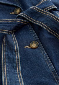 Tejido de mezclilla azul oscuro con costuras marrones, que presenta dos botones marrones y un detalle de tira o cinturón doblado.