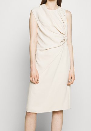 Ærmeløs midi-kjole i cremefarve med opsamlet knudedetalje i siden, båret af en stående person mod en ensfarvet baggrund.