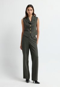 Zielony, dopasowany garnitur z kamizelką i spodniami z złotymi guzikami, o dopasowanym kroju i prostych nogawkach. Modelka nosi czarne, szpiczaste szpilki, neutralne tło.