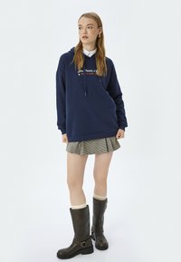Granatowy hoodie z sznurkami i haftowanym napisem, zestawiony z plisowaną spódnicą w kratkę oraz brązowymi botkami za kolano z paskami w okolicy kostki.