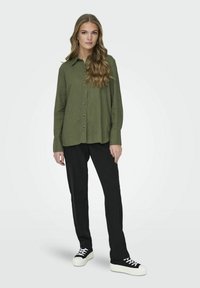 Olivgrünes Hemd mit Knopfleiste und Kragen, kombiniert mit schwarzen Hosen und schwarzen Sneakers mit weißen Sohlen. Glatter Stoff, lässige Passform.