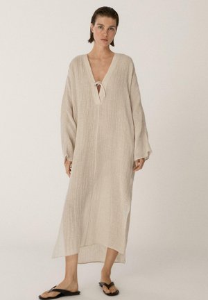 Frau trägt ein lockeres, langes beiges Kleid aus strukturiertem Stoff mit V-Ausschnitt und Bindeband, kombiniert mit schwarzen flachen Sandalen vor neutralem Hintergrund.