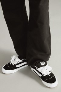 Zapatillas en blanco y negro con parte superior de ante, cordones blancos, tachuelas metálicas en la puntera y una etiqueta de logo en el talón. Suela de goma gruesa.