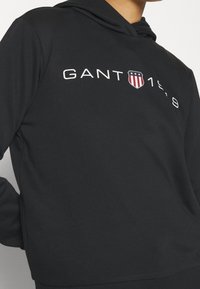 Černá mikina s kapucí vyrobená z jemného materiálu, na přední straně s grafikou "GANT 1949" v bílé barvě a s emblémem štítu s červenými a modrými akcenty.