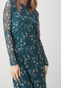 Abito a maniche lunghe color verde acqua con motivi floreali, caratterizzato da maniche trasparenti e scollo drappeggiato. Il tessuto ha una texture liscia.