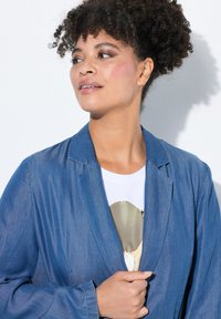 Denimblazer, ljusblå, enfärgad, med slagkrage, strukturerad tyg, lager på en vit topp med guldinslag.