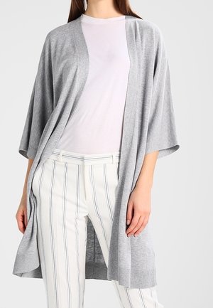 Frau trägt eine hellgraue, offen geschnittene Strickjacke über einem weißen T-Shirt und weißen Hosen mit vertikalen grauen Streifen.