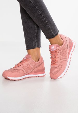 Personne portant des baskets New Balance 574 roses et un jean skinny noir retroussé, debout sur une surface blanche lisse.