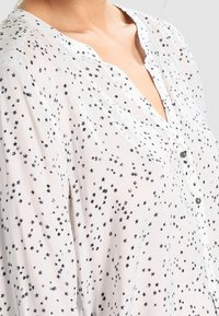 Camicia bianca trasparente con piccole stampe di stelle nere, chiusura con bottoni, scollatura a V e maniche larghe, che presenta una texture leggera.