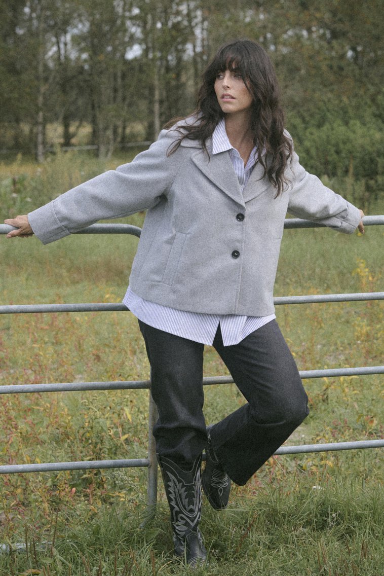 Giacca in misto lana grigia con grandi rever, due bottoni e tasche frontali, indossata sopra una camicia a righe e jeans neri, abbinata a stivali neri decorativi.