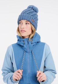 Berretto di lana blu con pompon e motivo a trama. Abbinato a una giacca blu chiaro con lacci regolabili.