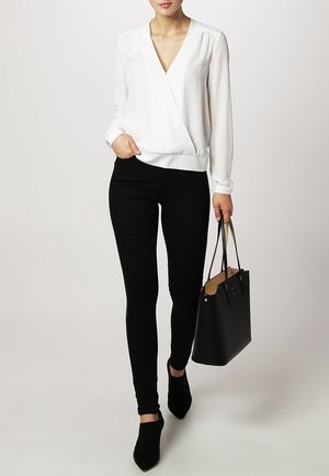 Blouse blanche à manches longues, assortie à un pantalon noir ajusté et des chaussures à talons noirs à bout pointu, tenant un sac fourre-tout noir.