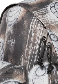 Reppu, jossa on teksturoitu musta ja beige kangas, jossa on abstrakteja kuvioita, kaareva muoto ja metallivetoketjut mustilla narunvetimillä.