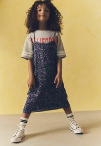 Blumenkleid in Marineblau mit lila und rosa Akzenten, ausgestattet mit dünnen Trägern, über einem grauen T-Shirt getragen, kombiniert mit weißen Turnschuhen und gestreiften Socken.