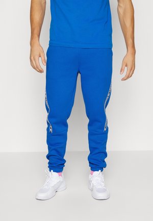 Siniset juoksuhousut, jotka on valmistettu pehmeästä kankaasta, ja joissa on kapeneva malli, joustavat hihnat ja valkoiset sivuviivat. Yhdistetty valkoisiin lenkkareihin.