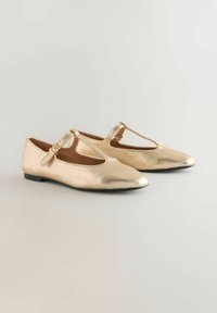 Par de zapatos planos metálicos dorados con tira en T y hebilla sobre un fondo neutro.