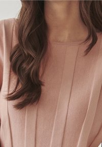 Měkký růžový svetr s ribbovanou strukturou, s vertikálními záhyby vpředu a kulatým výstřihem. Vlasy viditelné na jedné straně.