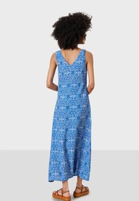 Vestido maxi azul con estampado, escote en V y tirantes anchos. Corte hasta el suelo, hecho de una tela ligera. Llevado con sandalias marrones.