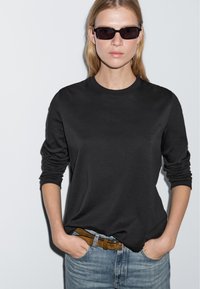 Schwarzes Langarmshirt aus weichem Stoff, mit Rundhalsausschnitt, kombiniert mit hellblauen Jeans und einem braunen Ledergürtel. Dunkle Sonnenbrille getragen.