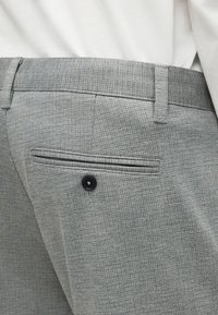 Κοντινό πλάνο γκρι υφασμάτινου παντελονιού με πίσω τσέπη welt ασφαλισμένη με μαύρο κουμπί, φορεμένο με λευκό πουκάμισο μπα tucked in.