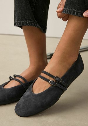 Black suede ballet flats featuring two narrow straps with metallic buckles, rounded toes, and a smooth, matte texture.
