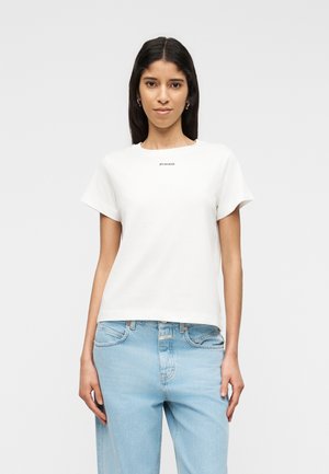 Hosszú sötét hajú nő fehér Pinko pólóban és világoskék farmert viselve, egyszerű világos háttér előtt áll.