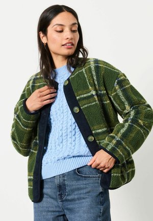 Kvinde iført blå strikket sweater, grøn ternet fleecejakke med knapper og blå jeans, poserer med den ene hånd, der holder jakke-kraven.