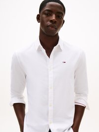 Camicia bianca con bottoni in tessuto leggero, dotata di un colletto classico, chiusura con bottoni e un logo discreto sul petto. Polsini piegati con accentuazione rossa.