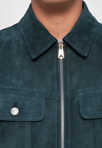 Veste en daim de couleur bleu sarcelle, dotée d'un col pointu, d'une fermeture éclair à l'avant avec quincaillerie dorée, et de deux poches poitrine avec pressions.