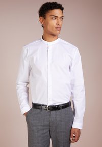 Camisa blanca de botones con cuello de banda, combinada con pantalones de cuadros grises y un cinturón negro. Tejido suave con mangas largas.