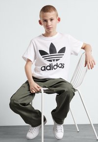 Balts kokvilnas T-krekls ar melnu Adidas logotipu, olīvzaļas krāsas kravas bikses un baltas kedas. Modelis sēž uz balta krēsla pret vieglu fonu.