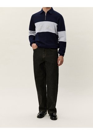 Hombre con polo de manga larga azul marino y gris con texto "Les Deux", vaqueros negros de corte holgado y zapatos negros brillantes, de pie con las manos en los bolsillos.