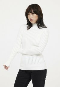 Weißer Strickpullover mit hohem Kragen, gerippter Textur und vertikalem Streifenmuster. Kombiniert mit schwarzen Jeans für einen Kontrast.