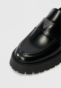 Black patent leather loafer with a smooth finish, stitched detailing, rubber sole, and a triangular metal logo accent on the upper strap.