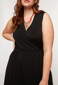 Svart ärmlös klänning med V-ringning, sammanknuten midja och en flödande kjol. Accessoarer i form av lager av halsband i guld- och rödtoner.