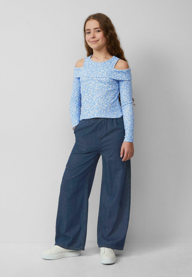 Hellblaues schulterfreies Oberteil mit weißem Blumenmuster, kombiniert mit locker sitzenden dunkelblauen Jeans und weißen Sneakers.