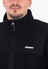Giacca in pile nera con zip e colletto alto. Presenta un mix di tessuti lisci e testurizzati, con un'etichetta con il marchio sul petto.
