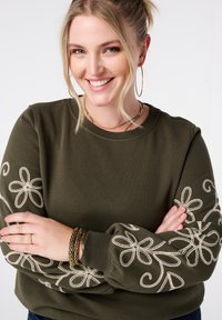 Olijfgroene sweatshirt met beige bloemborduurwerk op de mouwen. Zachte stof, ronde halslijn en een ontspannen pasvorm. Verstelbare manchetten.