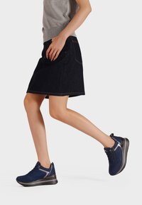 Donna che cammina con una gonna di jeans scuri, un maglione grigio e scarpe da ginnastica blu navy con suole spesse e dettagli argentati su sfondo bianco.