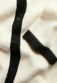 Mjuk krämfärgad cardigan med svart knapprem och svartkantade ärmslut, som visar upp ett mysigt texturerat tyg i närbild.