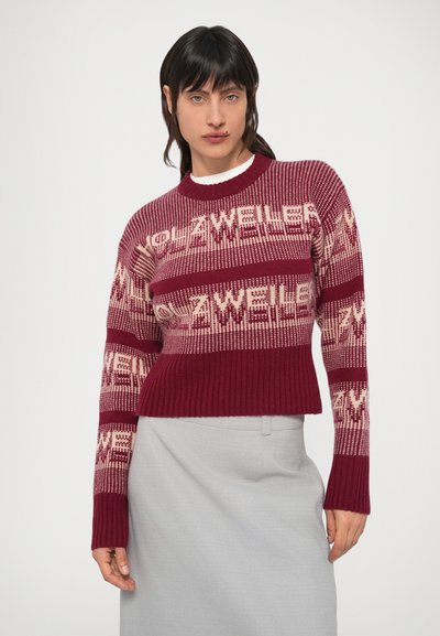 Vino rdeč pleten pulover s svetlo krem napisom v vzorcu, rebrastimi manšetami in skrajšano silhueto, nošen preko svetlo sive krila.