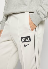 Nike sweatpants i ljusgrått med svarta detaljer, utrustade med sidofickor och en framträdande fyrkantig logotyplapp på låret. Mjuk textur.