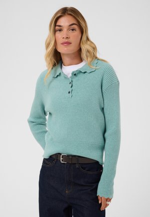 Femme aux cheveux blonds mi-longs portant un pull en tricot côtelé bleu clair par-dessus un haut blanc et un jean foncé avec une ceinture noire.