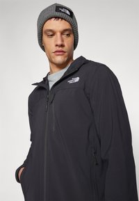 Schwarze, leichte Jacke mit Reißverschluss und Taschen; verfügt über eine Kapuze und das Logo von The North Face auf der Brust. Getragen mit einer grauen Strickmütze.