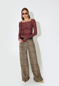 Bordeauxfarbenes Spitzenoberteil mit langen Ärmeln, kombiniert mit weiten Leopardenmuster-Hosen mit hoher Taille und spitzen goldenen Schuhen. Das Model trägt eine dunkle Sonnenbrille.
