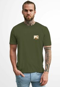 Olive green short-sleeve t-shirt with a chest pocket graphic featuring mountains and a sun in beige and orange colors. Cotton fabric, plain texture.