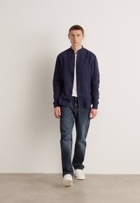 Marineblå linbutton-up-skjorte, hvit crew neck t-skjorte under, mørke jeans og hvite joggesko. Enkel design og teksturert stoff.