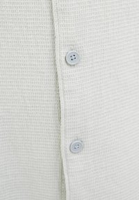 Camisa de botones de tela texturizada de color gris claro con un patrón de cuadrícula, que presenta botones redondeados en gris a lo largo del frontal.