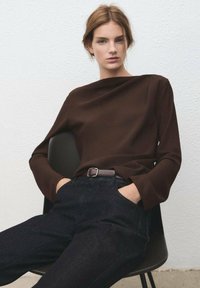 Hnedý top s dlhým rukávom a výstrihom loďkového tvaru, v kombinácii s tmavými džínsami a hnedým opaskom. Hladká textúra, priliehavý strih, minimalistický štýl.